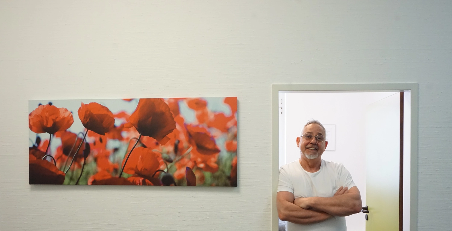 Praxis für Chiropraktik - Therapien und Behandlungs -  Henry Burghammer steht mit verschränkten Armen in einem weißen T-Shirt in einem Türrahmen neben einem großen Panorama-Wandbild mit leuchtend roten Mohnblumen.